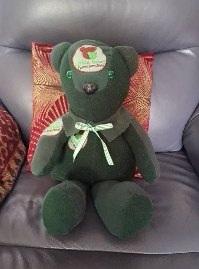 A memory bear made from a dark green fleece with an embroidered preschool logo on its head, finished with a striped ribbon bow.